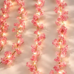 Peut inclure: Une guirlande de fleurs de cerisier artificielles dans des tons roses, intégrant des lumières LED blanc chaud. La guirlande florale se compose de multiples pétales délicats disposés le long d'un fil flexible, créant un effet lumineux.
