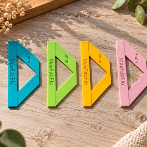 Könnte beinhalten: Vier dreieckige Nahtführungen in Blau, Grün, Gelb und Pink. Jede Führung ist mit dem Text MaxFabFlo und einem spezifischen Maß von 2mm, 3mm, 4mm oder 5mm bedruckt. Diese Werkzeuge sind für das Nähen und die Stoffausrichtung konzipiert.