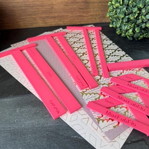 Könnte beinhalten: Helle pinkfarbene MaxFabFlo-Werkzeuge mit Millimeter-Messungen liegen auf gemustertem Papier. Die Werkzeuge sind wahrscheinlich für Bastel- oder DIY-Projekte gedacht. Der Hintergrund zeigt dekoratives Papier und eine Holzoberfläche.