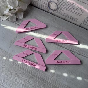 Könnte beinhalten: Fünf rosa MaxFabFlo-Werkzeuge mit dreieckigem Design. Die Werkzeuge sind mit den Maßen 3 mm und 4 mm beschriftet. Die Werkzeuge sind auf einer hellen Holzoberfläche angeordnet.