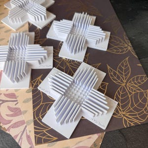 Könnte beinhalten: Weiße, kreuzförmige Objekte mit einem geschichteten Design werden auf gemustertem Papier präsentiert. Die Kreuze haben ein strukturiertes, geometrisches Aussehen. Der Hintergrund zeigt verschiedene Papierdesigns mit Blumen- und Blättermustern in Braun-, Beige- und Lilatönen.