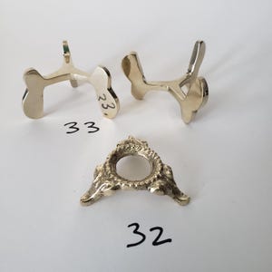 May include: Three metallic display stands with gold-toned finishes. Two stands feature smooth, curved tripod designs labeled with the number 33. One stand has a textured, circular base with a central opening and is labeled with the number 32.