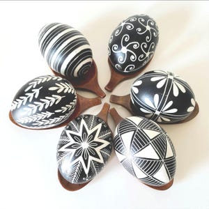 May include: Six decorative Easter eggs with intricate black and white designs. Each egg rests on a small wooden spoon. The eggs feature various patterns, including stripes, floral, and geometric shapes.