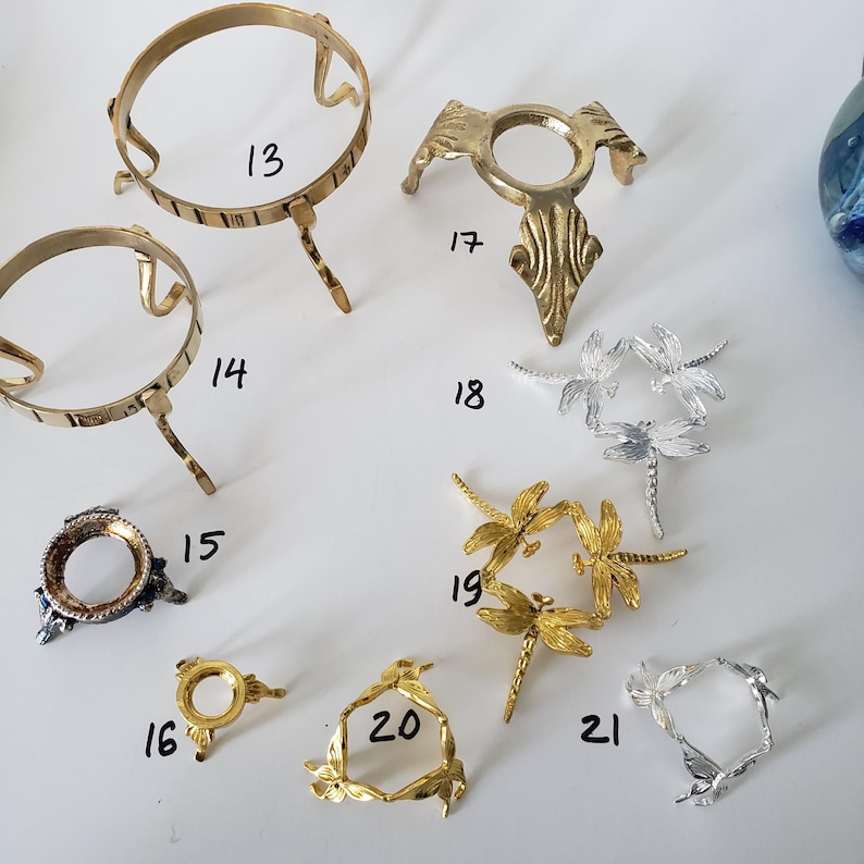 May include: A collection of metal display stands labeled 13 through 21. The assortment includes gold-toned circular wire stands, a decorative brass tripod stand, silver and gold dragonfly-shaped stands, and small ornate metal bases for spheres.