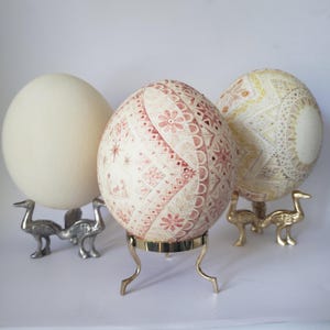 Peut inclure: Trois grands œufs de forme ovale présentés sur des supports en métal. L'œuf de gauche est crème uni. L'œuf central présente des motifs géométriques et floraux rouges complexes. L'œuf de droite affiche des motifs étoilés jaunes et dorés.