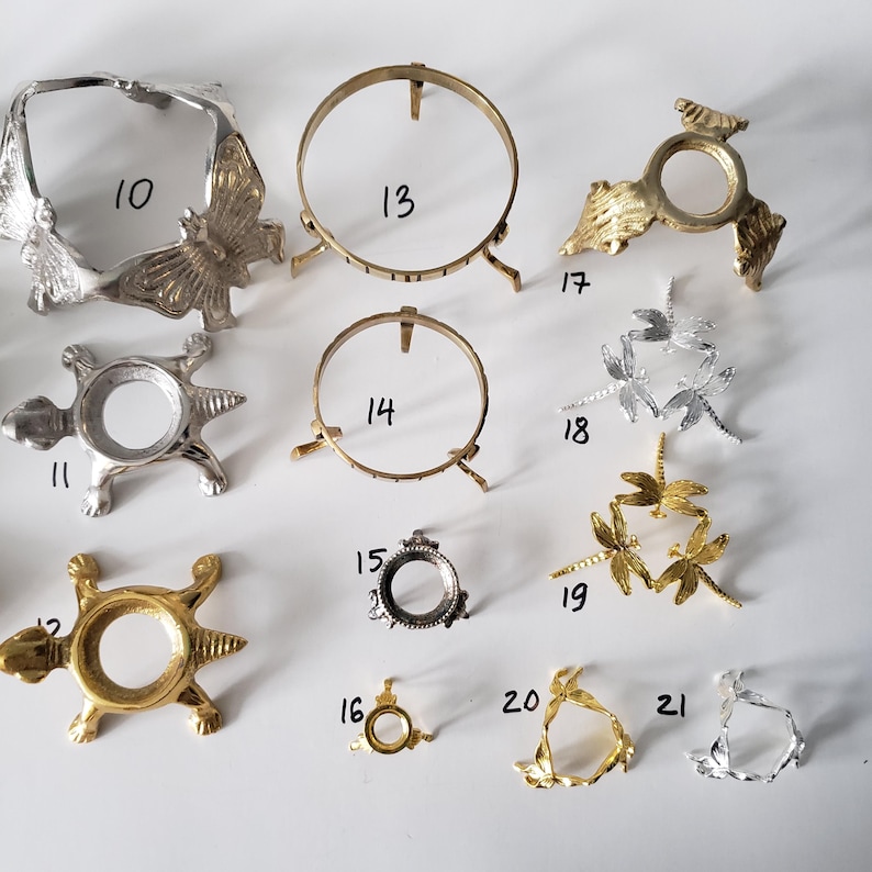 May include: A collection of twelve metal display stands in silver and gold tones, labeled 10 through 21. The stands feature various designs including butterflies, turtles, dragonflies, and simple rings, intended for holding spheres or small objects.