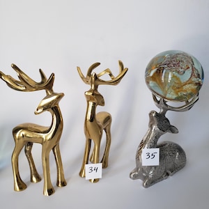 May include: Three deer figurines: two standing brass deer with polished gold-tone finishes and one silver-tone metal deer in a seated position, balancing a round, multi-colored glass orb on its antlers. A white tag labeled 34 and 35 is attached.