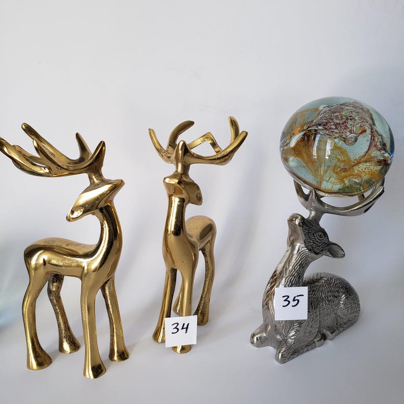 May include: Three deer figurines: two standing brass deer with polished gold-tone finishes and one silver-tone metal deer in a seated position, balancing a round, multi-colored glass orb on its antlers. A white tag labeled 34 and 35 is attached.