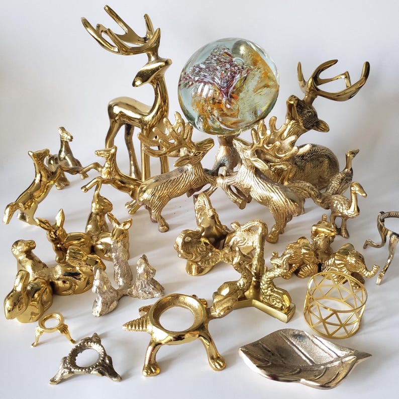 May include: A collection of polished brass animal figurines including several deer, elephants, and birds, alongside various metal display stands, a leaf-shaped dish, and a clear glass sphere with internal colorful inclusions and air bubbles.