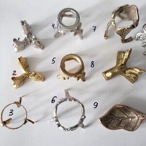 May include: A collection of nine metal display stands and holders in various shapes and finishes, including silver and gold tones. The items are numbered 2 through 9, featuring tripod bases, ring-shaped tops, and a leaf-shaped tray, all arranged on a white surface.