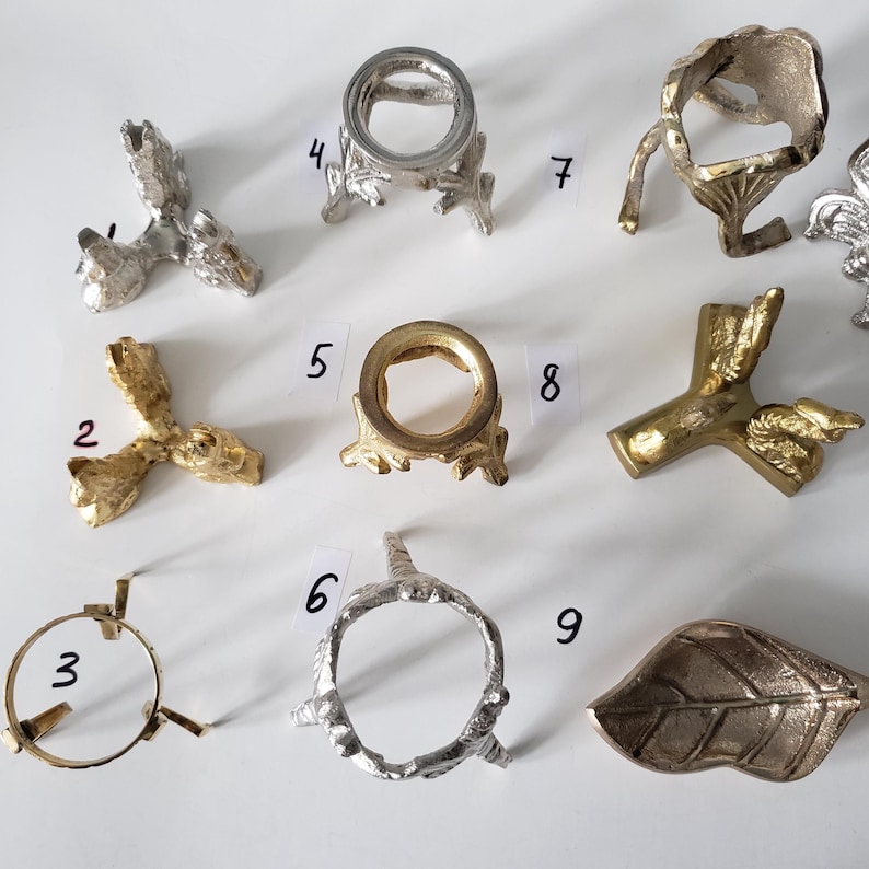 May include: A collection of nine metal display stands and holders in various shapes and finishes, including silver and gold tones. The items are numbered 2 through 9, featuring tripod bases, ring-shaped tops, and a leaf-shaped tray, all arranged on a white surface.