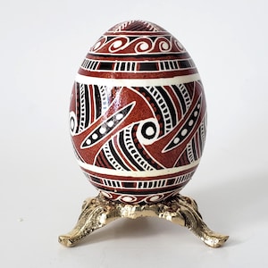 May include: A decorative, hand-painted egg with a brown base color, featuring intricate patterns in white and black. The egg is displayed on a gold-colored, ornate stand, showcasing its detailed design and craftsmanship. A unique Easter or home decor item.