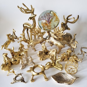 May include: A selection of gold-toned decorative ornaments and objects. The display features deer, bird figurines, and other animal sculptures, alongside decorative stands and a leaf-shaped dish. A glass orb with colourful interior details is also included.