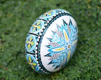 May include: A white egg with intricate, hand-painted designs in blue, yellow, and black. The egg features a floral pattern on one side and geometric shapes on the other. The egg is resting on a bed of green grass.