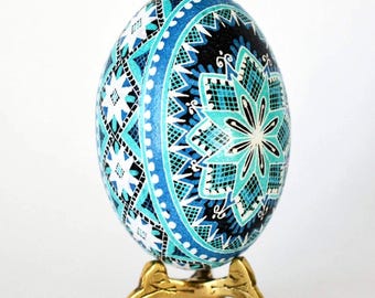 May include: A decorative egg with a detailed geometric design in shades of blue, turquoise, and white, set against a black background. The egg is displayed on a small, ornate gold-colored stand. The egg's pattern includes star and floral motifs.
