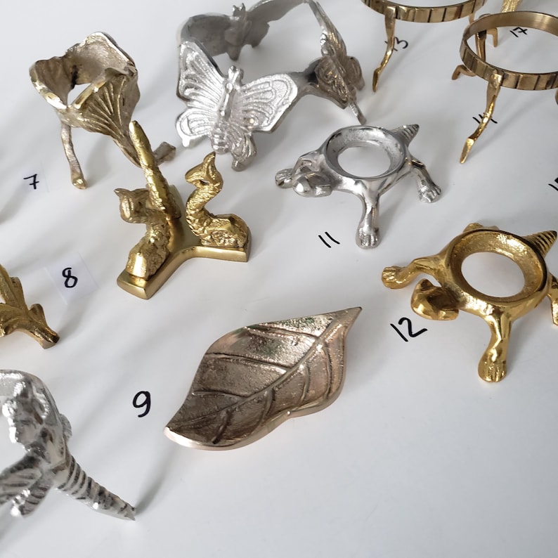 May include: A collection of metal display stands and holders in gold and silver tones, featuring various shapes including a butterfly, a leaf, and animal-inspired designs. Each piece is labeled with a small black number for identification purposes.