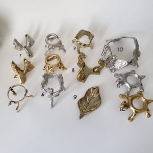 May include: A collection of twelve metal display stands and holders in silver and gold tones, featuring various designs including butterflies, turtles, and leaf shapes. Each item is numbered from 1 to 12, showcasing different structural forms.