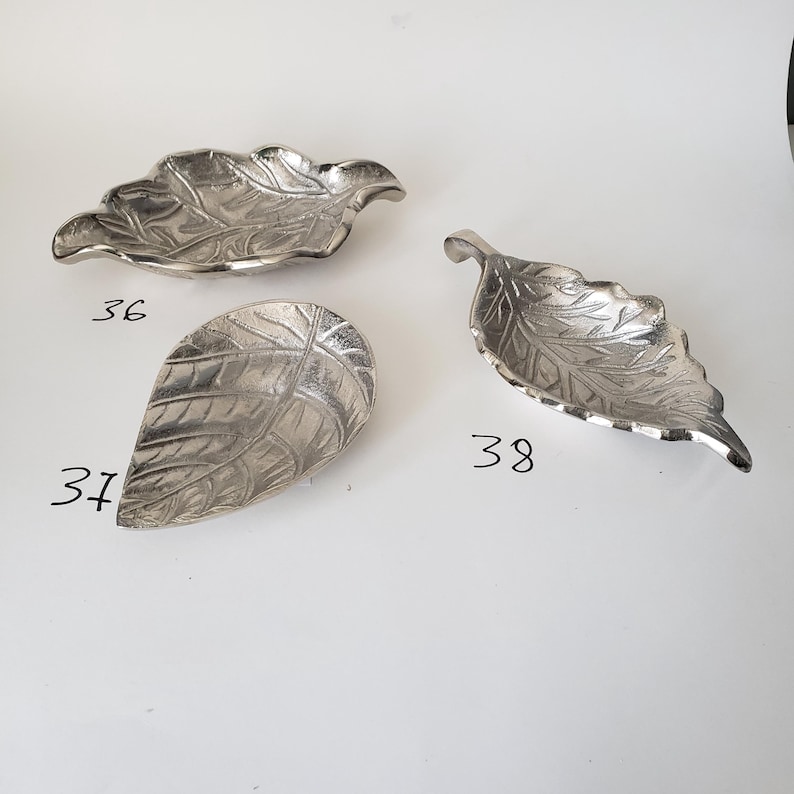 May include: Three silver-colored metal dishes shaped like leaves, each with detailed vein textures. The dishes are labeled with the numbers 36, 37, and 38 written in black ink on the surface, identifying three distinct leaf-shaped designs.