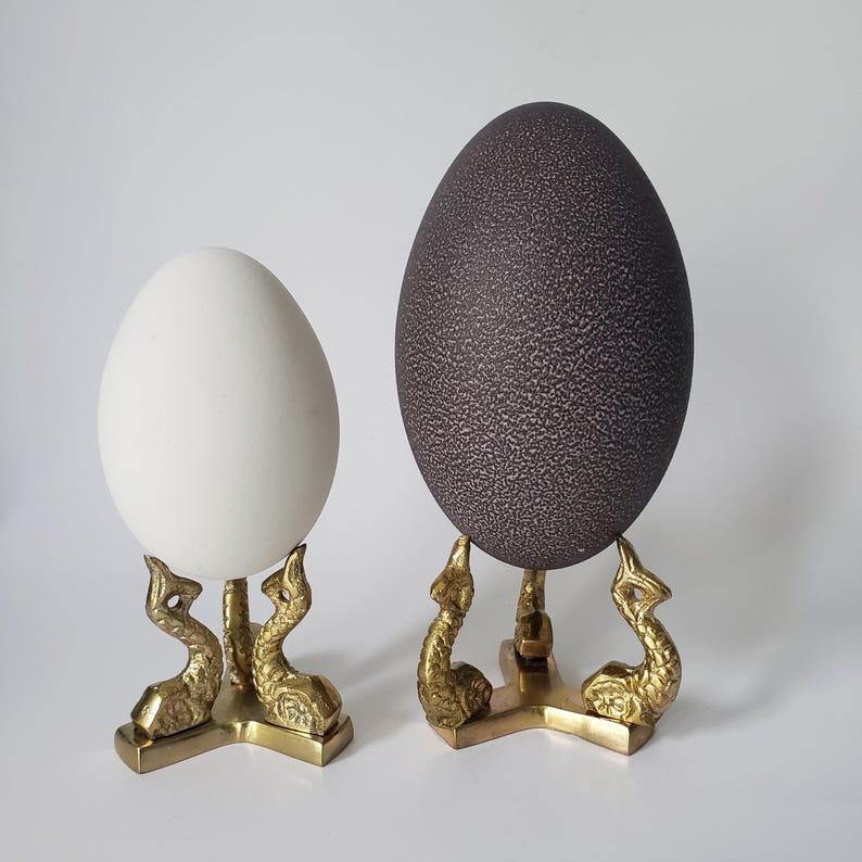 May include: Two oval-shaped decorative eggs displayed on gold-toned metal stands. One egg is smooth and white, while the larger egg is dark brown with a textured, speckled surface. Each stand features three legs shaped like stylized sea creatures.