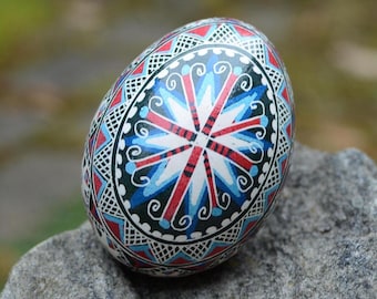 Op de afbeelding: Een versierd ei met een kleurrijk ontwerp. Het ei heeft een centraal sterpatroon in rood, wit en blauw, omringd door ingewikkelde geometrische en bloemmotieven. Het ei ligt op een grijze steen.