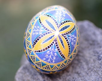 May include: A hand-painted Easter egg with a blue, yellow, and black geometric design. The egg features a stylized flower pattern in the center.