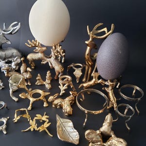 Puede incluir: Una colección de soportes y expositores decorativos para huevos en varios diseños y acabados metálicos. La imagen presenta un huevo grande de color crema y un huevo gris oscuro, cada uno exhibido en soportes ornamentados. Otros soportes incluyen diseños de animales y hojas.