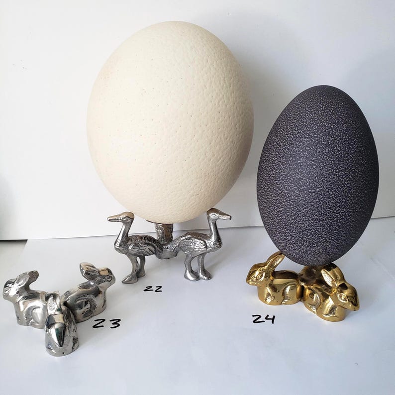 May include: A large cream-colored egg on a silver-toned bird-shaped stand labeled 22, a silver-toned rabbit-shaped stand labeled 23, and a dark gray egg on a gold-toned rabbit-shaped stand labeled 24, all arranged on a white surface.