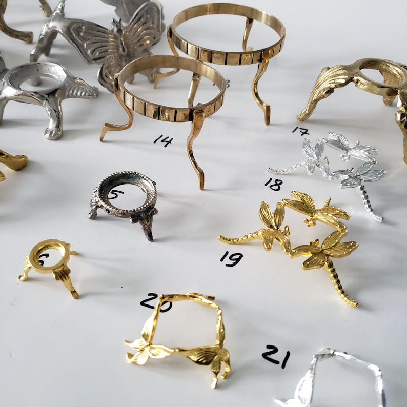 May include: A collection of metal display stands in gold and silver tones, featuring various designs including butterflies, dragonflies, and simple tripod bases. Each stand is labeled with a handwritten number for identification and organization.
