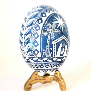 May include: A blue and white egg ornament featuring intricate hand-painted details including a nativity scene with a star, palm trees, and geometric patterns. The egg is displayed on a small, three-legged gold-toned metal stand.