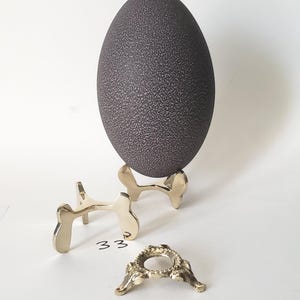 May include: A dark brown, textured emu egg resting on a polished gold-tone metal stand. Beside it sits an additional gold-tone metal stand labeled 33 and a smaller, ornate gold-tone metal ring stand labeled 32, both featuring metallic finishes.