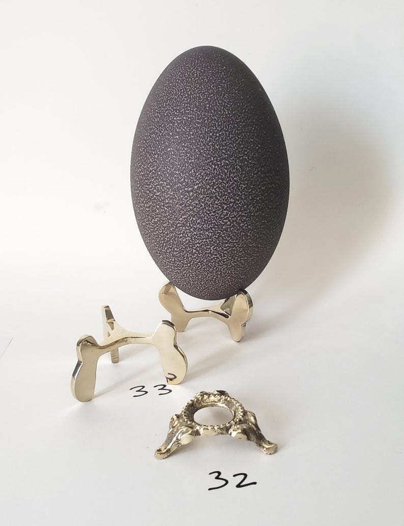 May include: A dark brown, textured emu egg resting on a polished gold-tone metal stand. Beside it sits an additional gold-tone metal stand labeled 33 and a smaller, ornate gold-tone metal ring stand labeled 32, both featuring metallic finishes.