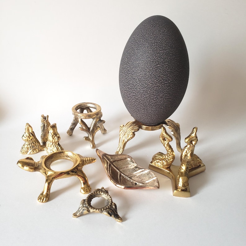 May include: A dark gray textured egg sits on a gold-toned metal stand with leaf-shaped feet. Surrounding it are several brass-colored metal display stands, including one shaped like a turtle, one with bird figures, and a small leaf-shaped dish.