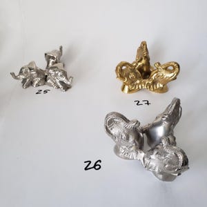May include: Three metal elephant-shaped stands arranged in a triangular formation. Item 25 is silver-toned, item 26 is a larger silver-toned version, and item 27 is gold-toned. Each stand features three elephant heads facing outward.