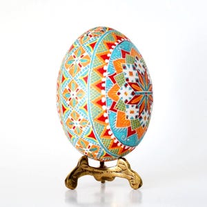 May include: A vibrantly decorated egg with intricate geometric patterns in red, orange, blue, and white, displayed on a small, ornate gold-colored stand. The egg's design features floral and star-like motifs, creating a festive and artistic appearance.