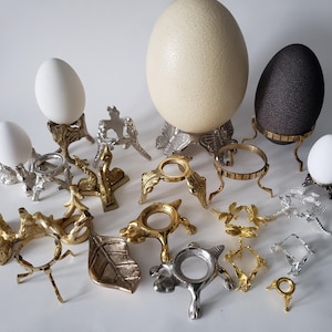 May include: An assortment of decorative egg holders in various metallic finishes, including gold and silver. The holders are designed with ornate, nature-inspired details, such as leaves and dragonflies. Several eggs, including a large ostrich egg, are displayed in the holders.