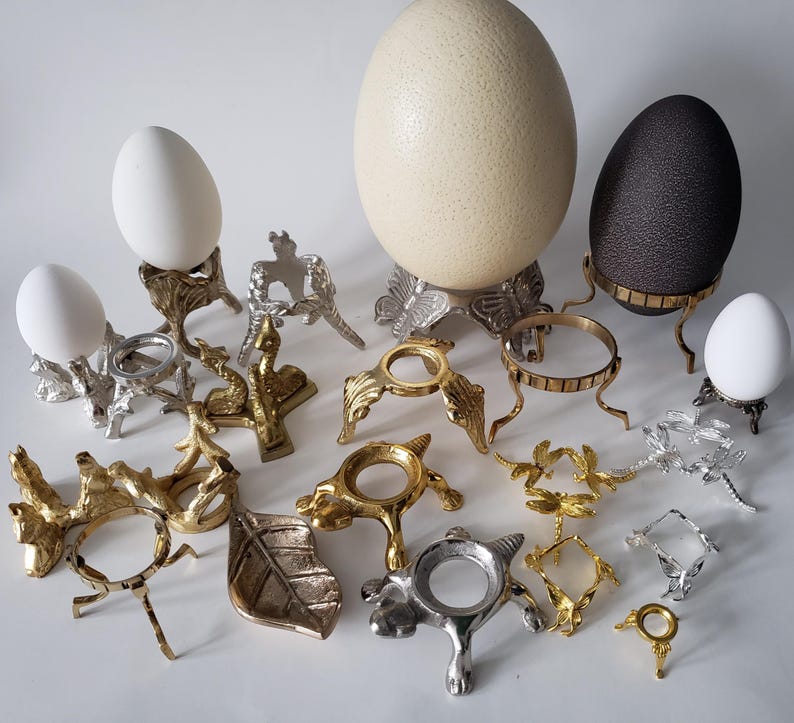 May include: A collection of decorative metal egg stands in various finishes including gold, silver, and bronze. The stands feature intricate designs such as butterflies, turtles, and leaves. Several white eggs and one large cream and dark brown egg are displayed.