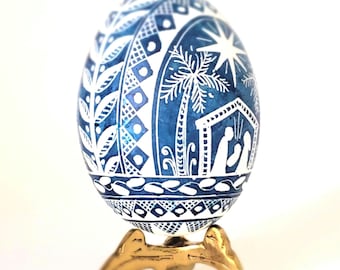 May include: A blue and white decorative egg with a religious theme, displayed on a gold-coloured stand. The egg features intricate white patterns and a nativity scene with a star and palm tree.