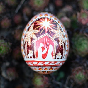 Puede incluir: Un huevo de Pascua decorado en rojo, blanco y dorado con una escena de la Natividad pintada en él. El huevo presenta una estrella, palmeras y un pesebre con María, José y el niño Jesús.