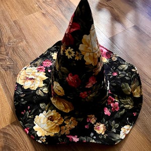 May include: A black witch hat with a pointed crown and a wide brim. The hat is covered in a floral print with pink, yellow, and red flowers on a black background.