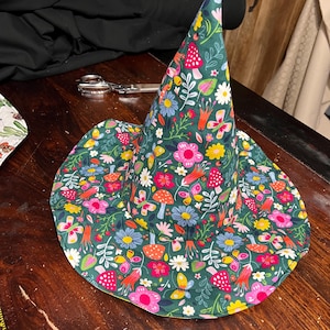 May include: A whimsical witch hat with a wide brim, featuring a vibrant floral and mushroom pattern on a dark green background. The hat's design includes pink, yellow, and blue flowers, along with orange mushrooms and butterflies.