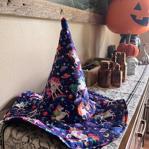 May include: A whimsical, quilted witch hat with a wide brim, featuring a vibrant print of unicorns, rainbows, planets, and stars on a deep blue background. The hat has a pointed top and is designed for Halloween or costume use.