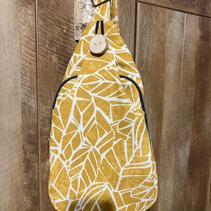 May include: A mustard yellow crossbody bag with a white geometric leaf pattern. The bag has a black strap and a wooden button closure.