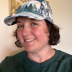 May include: A baseball cap with a forest and owl design. The cap features a forest scene with evergreen trees and a blue sky on the front panel. The side panels display a pattern of owls. The brim is a light blue and white pattern.