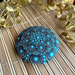 Puede incluir: Una piedra redonda pintada de azul y blanco con un diseño de mandala. La piedra está sobre una estera de bambú tejida.
