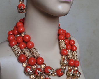 May include: A three-strand necklace and matching earrings set. The necklace features large, round, red beads interspersed with gold-colored filigree and smaller, clear beads. The earrings have a similar design, with a gold-colored top and a red bead.