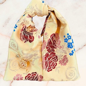 May include: A pale yellow fabric pouch with a knotted top handle, featuring a woven floral pattern in shades of deep red, pink, and bright blue. A gold-toned safety pin with a small, intricate, spherical filigree charm is attached to the front.