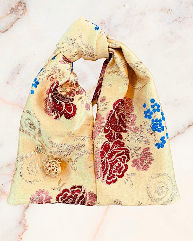 May include: A pale yellow fabric pouch with a knotted top handle, featuring a woven floral pattern in shades of deep red, pink, and bright blue. A gold-toned safety pin with a small, intricate, spherical filigree charm is attached to the front.