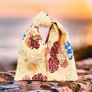 May include: A pale yellow fabric bag with a knotted handle, featuring a woven floral pattern in shades of deep red, pink, and bright blue. A small gold-toned safety pin with a decorative filigree charm is attached to the front of the bag.