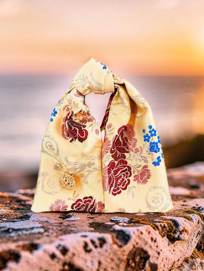 May include: A pale yellow fabric bag with a knotted handle, featuring a woven floral pattern in shades of deep red, pink, and bright blue. A small gold-toned safety pin with a decorative filigree charm is attached to the front of the bag.