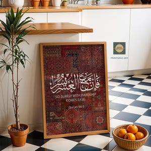 Pu&ograve; includere: Stampa incorniciata con un motivo a tappeto rosso e marrone, calligrafia araba e la frase inglese "SO, SURELY WITH HARDSHIP COMES EASE". L'opera d'arte &egrave; esposta in una cucina, insieme a una pianta in vaso e un cesto di arance.